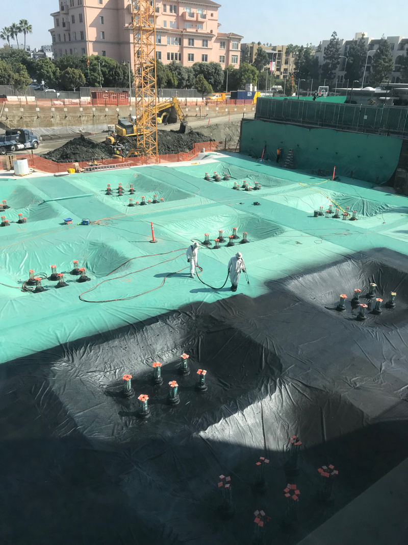 workers applying waterproof materials to a large area of ground in preparation for a new construction project to be built on that foundation.