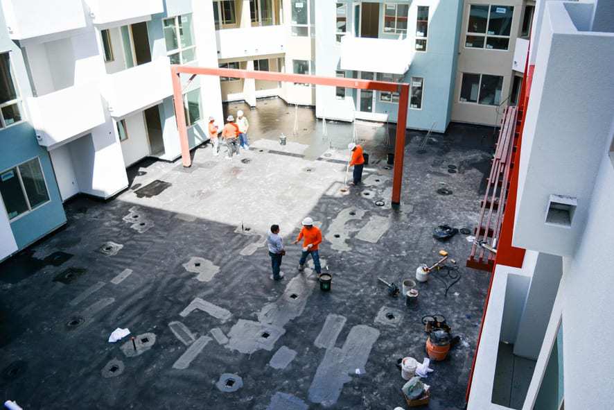 This job site is in the middle of various buildings. There’s an orange bar-like structure on the site, and there are people wearing orange shirts and hard hats on the site.