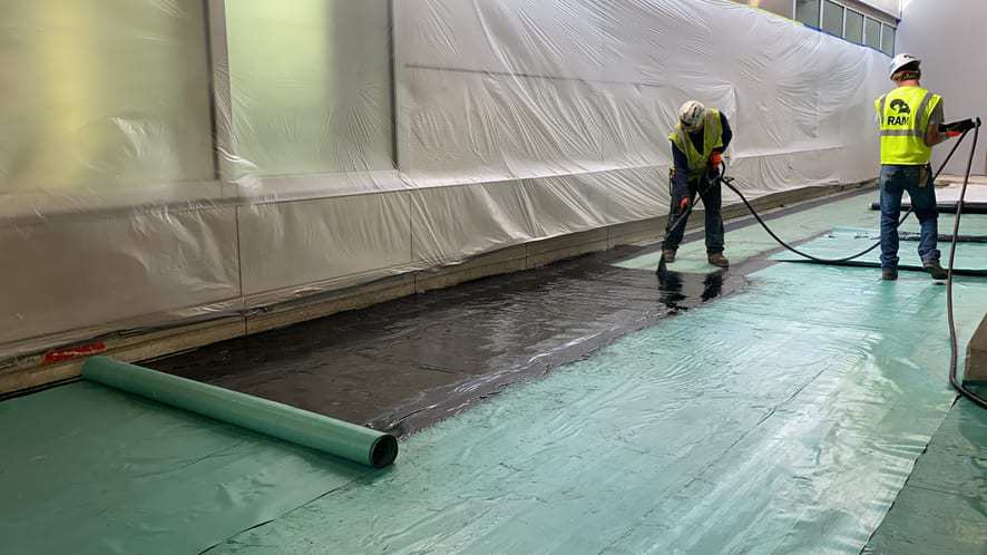 Two people are applying a waterproofing solution to a job site. They are spraying a black liquid to a green surface. They are wearing neon yellow safety vests and white hard hats.
