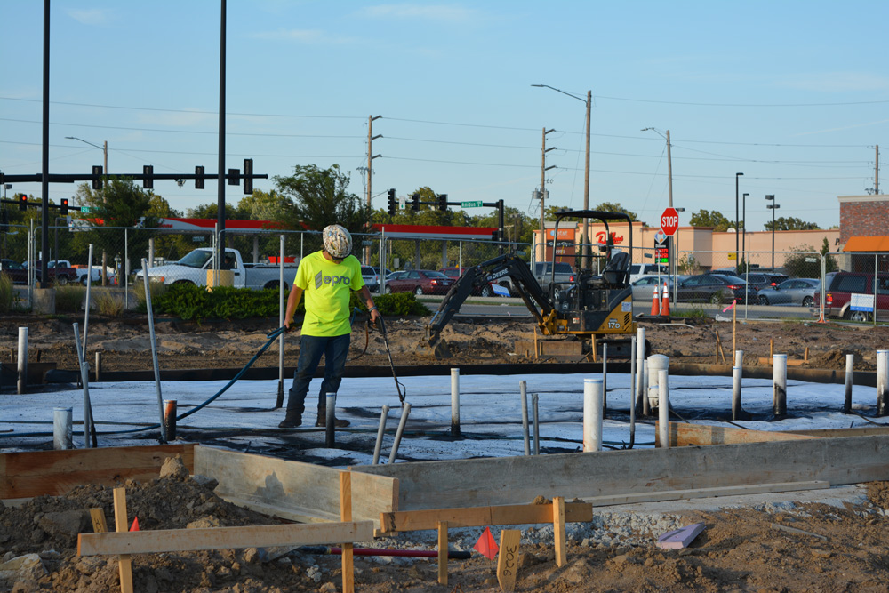 A person is applying a waterproofing solution to a job site. They are wearing a neon yellow shirt with dark jeans and a hard hat.